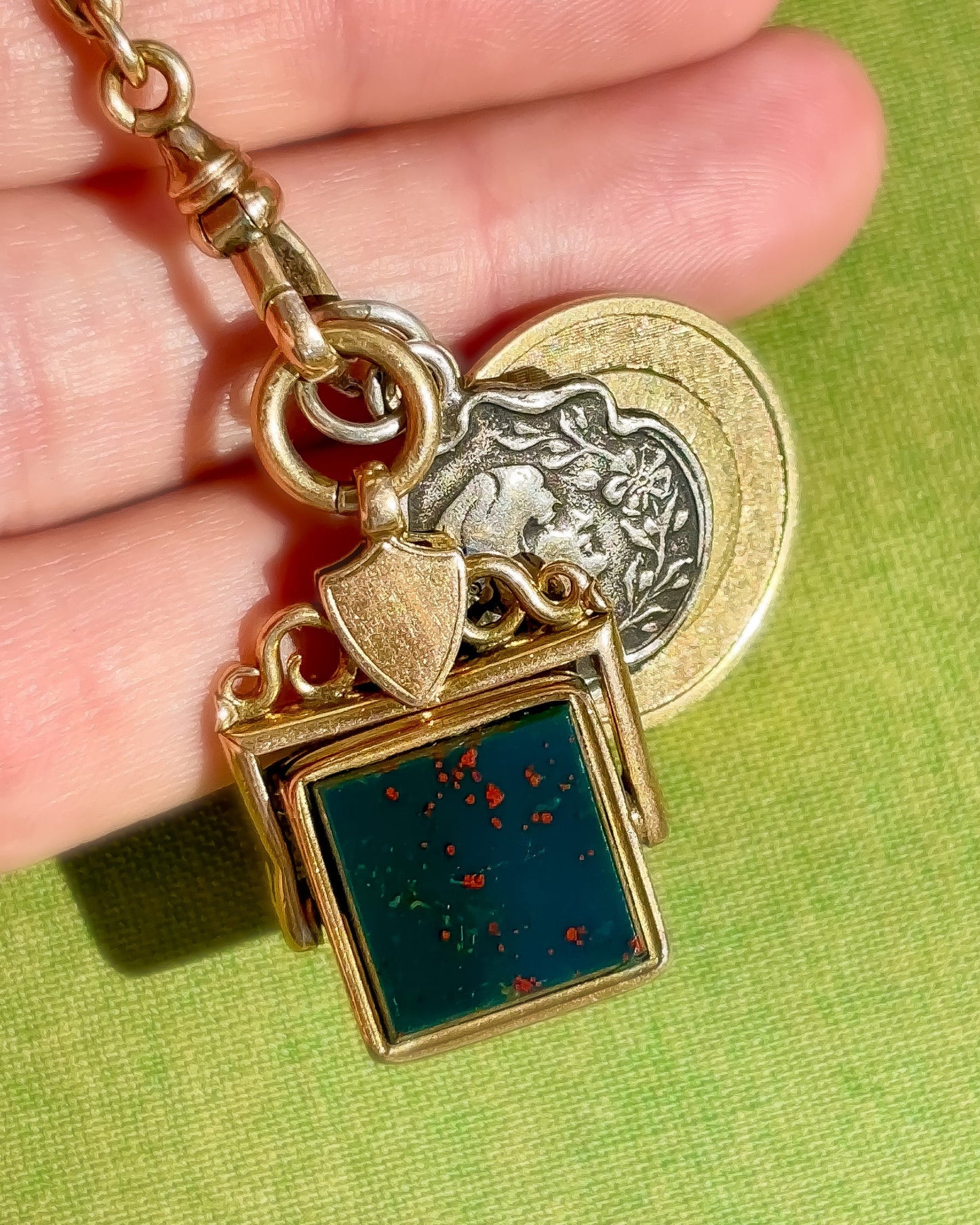 Antique gold fob pendant with a green bloodstone gemstone and decorative charms held in a hand against a green background