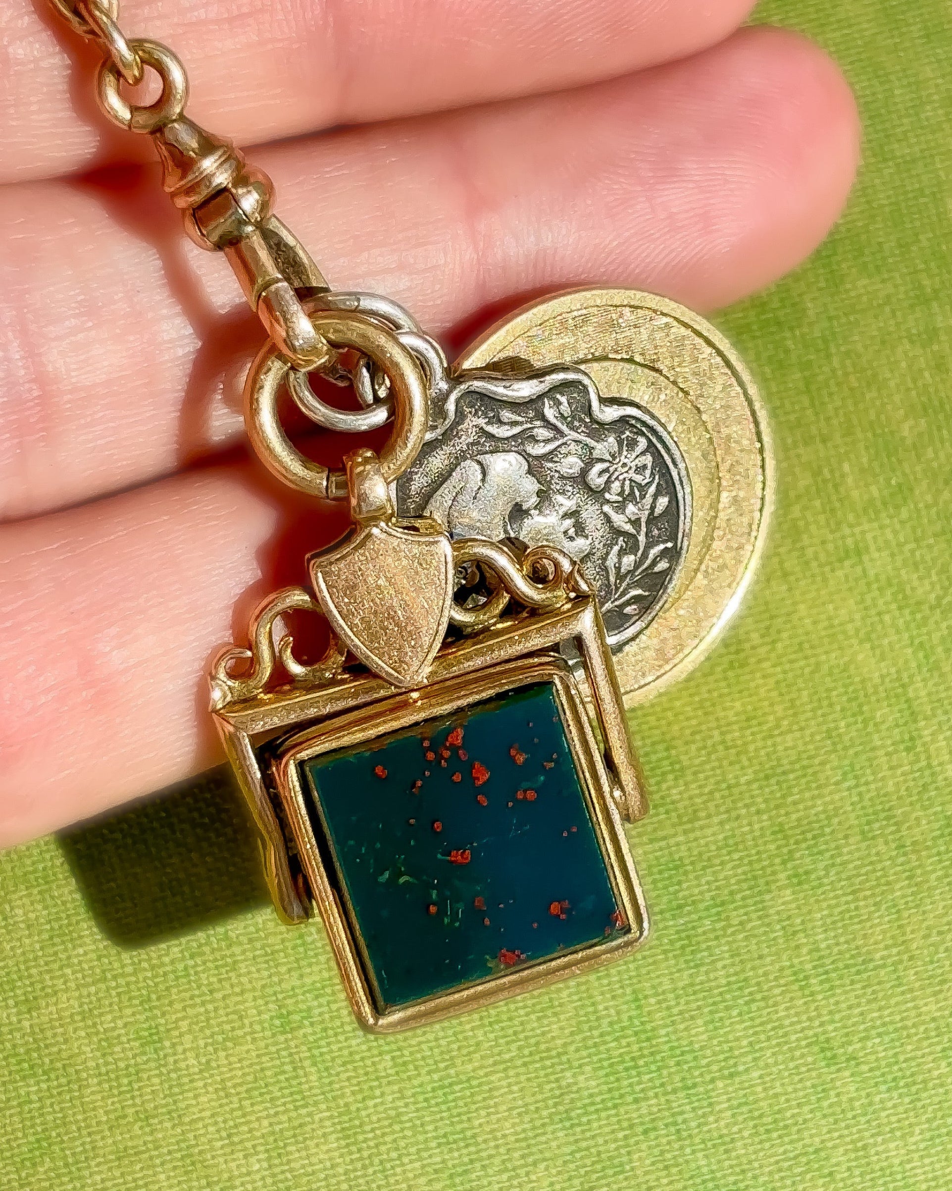 Antique gold fob pendant with a green bloodstone gemstone and decorative charms held in a hand against a green background